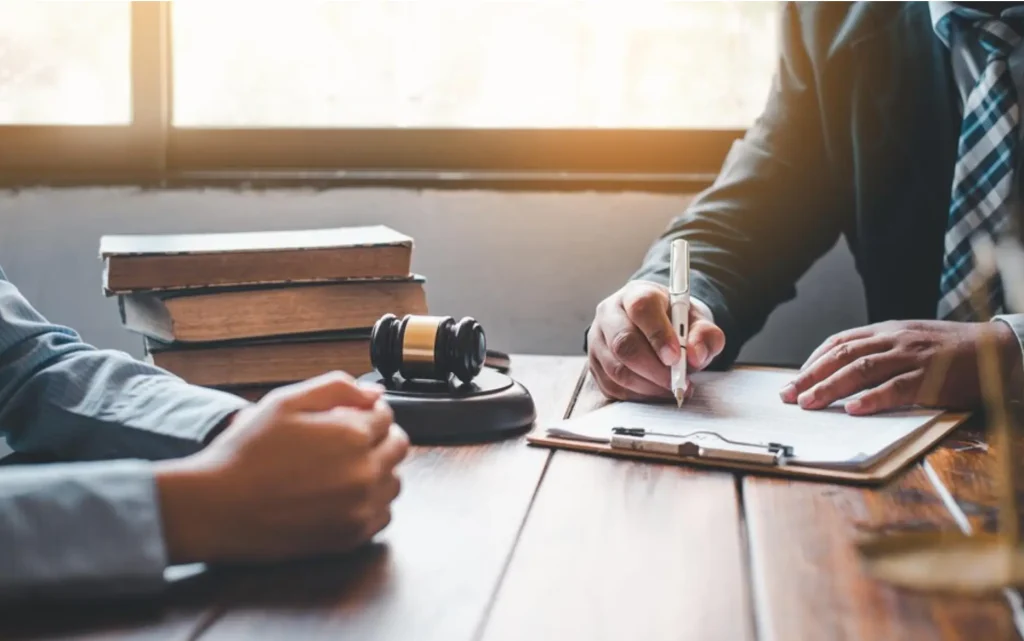 A person signing a document on a wooden desk, with a gavel and legal books in the background, suggesting a legal consultation or agreement.
