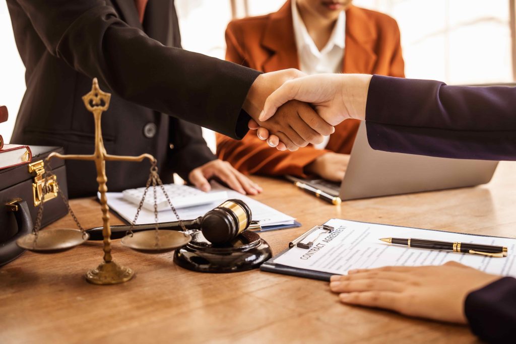 Lawyers and clients shaking hands over a contract agreement with gavel and justice scales on the desk.