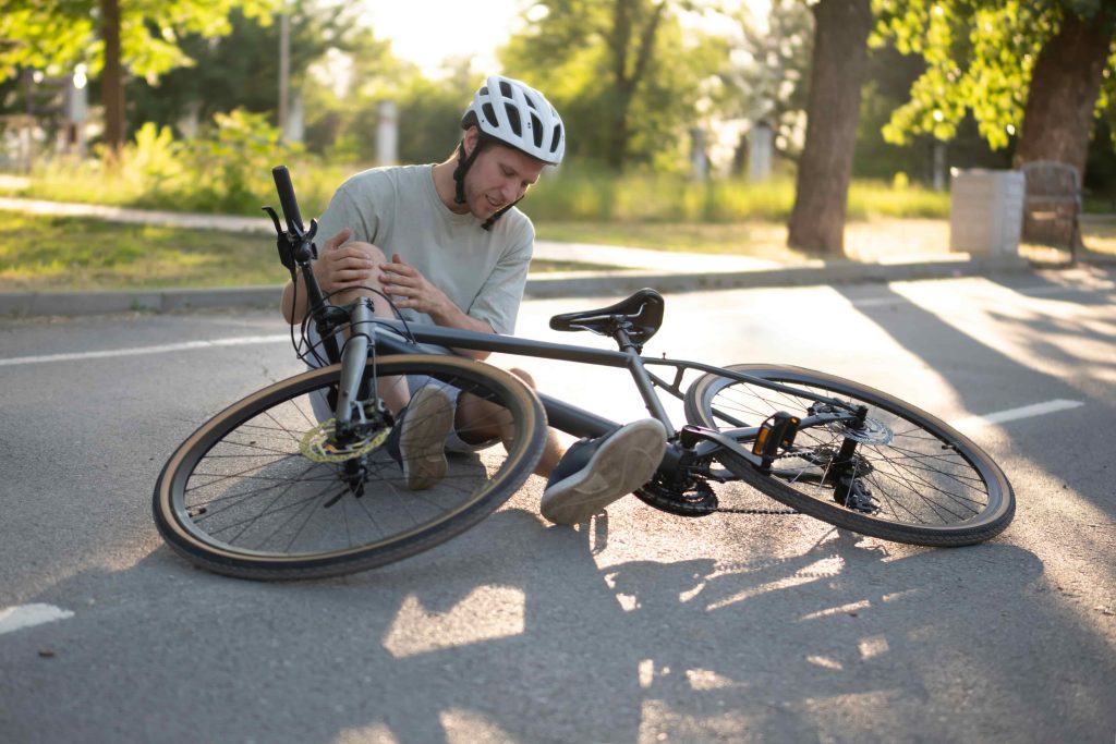 Cyclist wearing helmet injured after falling off bicycle on the road, holding his knee in pain.