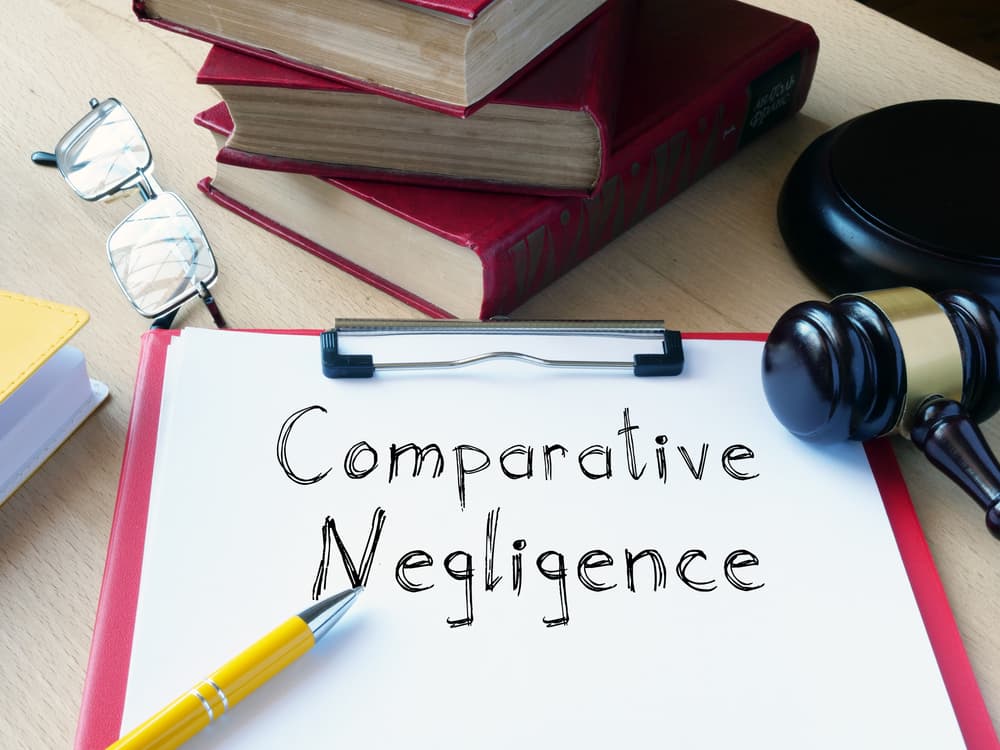 Clipboard with the words “Comparative Negligence” written on paper, placed on a desk alongside legal books, a judge’s gavel, eyeglasses, and a yellow pen, suggesting a legal or personal injury context.