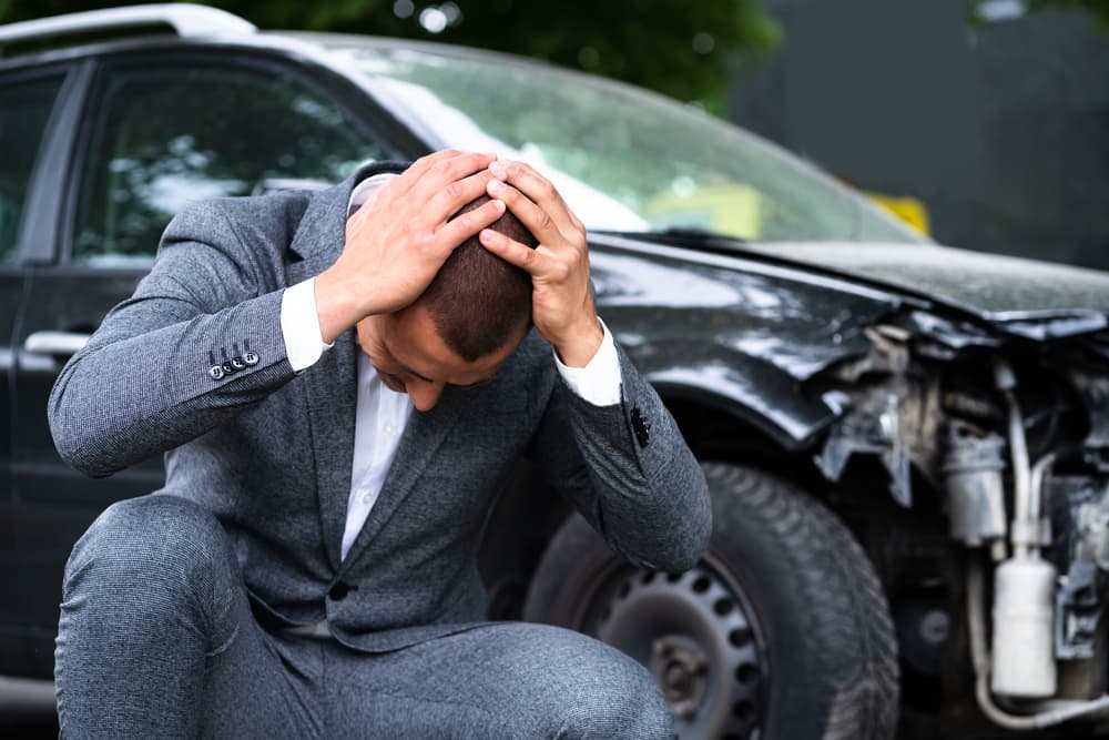 A man in a gray suit sits beside his damaged car, holding his head in pain and distress, symbolizing emotional suffering after a personal injury accident.
