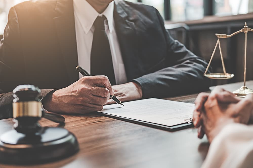 A lawyer in a suit sitting at a wooden desk, signing legal documents during a meeting with a client, with a gavel and scales of justice placed nearby.
