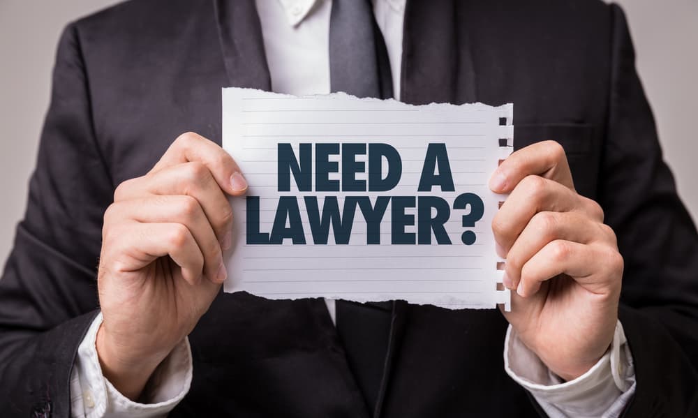 A man in a business suit holds a torn piece of paper with the bold text “NEED A LAWYER?” printed on it, symbolizing legal help or consultation.