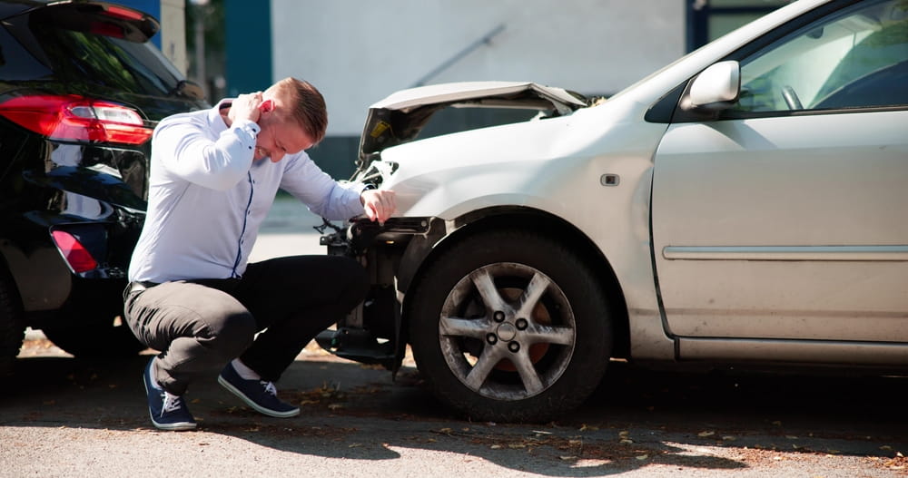 Conductor inspeccionando los da&ntilde;os en la parte delantera de un autom&oacute;vil despu&eacute;s de un accidente